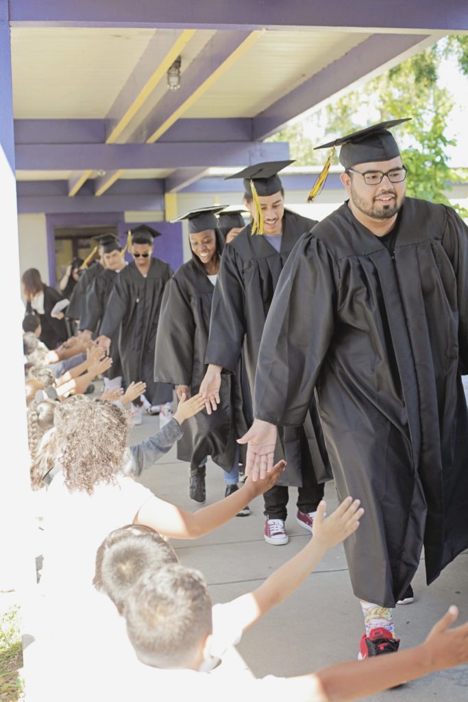 Graduates in cap & gown walking in a line and high-fiving young students as they walk by.