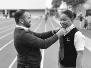 AGA Principal Cabrera placing a tie onto scholar, photo is in black and white and takes place outside.