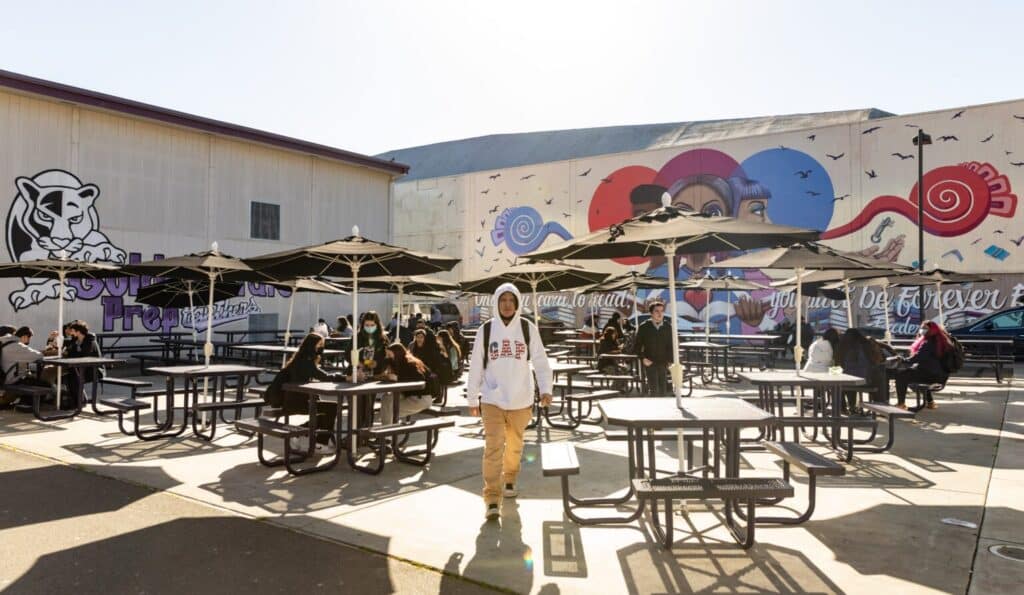 Campus shot of Aspire Golden State Prep with a high schooler walking through the quad with tables and murals behind him.
